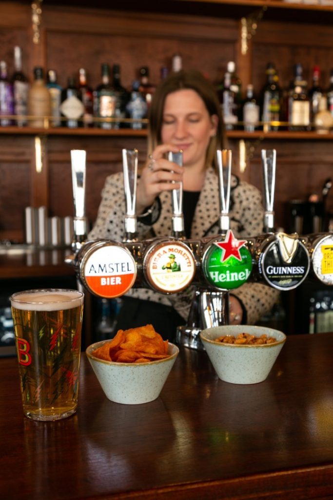 Pint of beer beside bar snacks with beer taps in the background